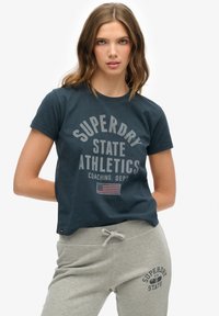 Mørkeblå bomulds t-shirt med korte ærmer, der har et cirkelskilt i lys grå og et lille flag detalje. Sammen med grå sweatpants.