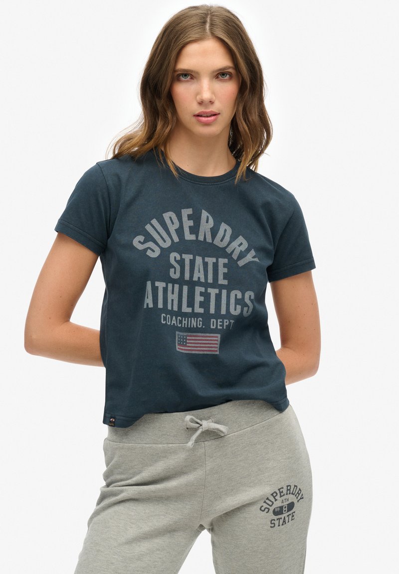 Mørkeblå bomulds t-shirt med korte ærmer, der har et cirkelskilt i lys grå og et lille flag detalje. Sammen med grå sweatpants.