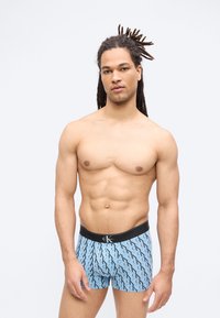 Άνδρας χωρίς πουκάμισο με πλεξίδες, φοράει ανοιχτό μπλε μποξέρ Calvin Klein με μαύρο σχέδιο λογότυπου, στέκεται σε απλό φόντο.