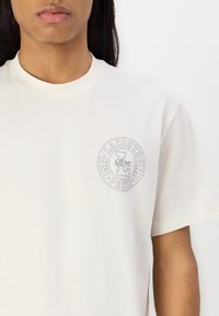 Cămașă T-shirt din bumbac light cream cu un logo brodat circular, cu inscripțiile "LACOSTE" și "PARIS", înconjurat de frunze de laur. Mâneci scurte.