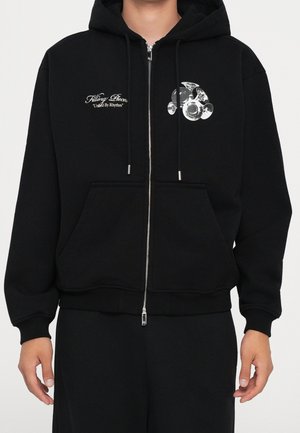 Czarna bluza z kapturem na zamek błyskawiczny z kieszeniami z przodu, biała grafika grupy trąbek na lewej stronie klatki piersiowej oraz tekst „Filling Pieces United By Rhythm” na prawej stronie klatki piersiowej.