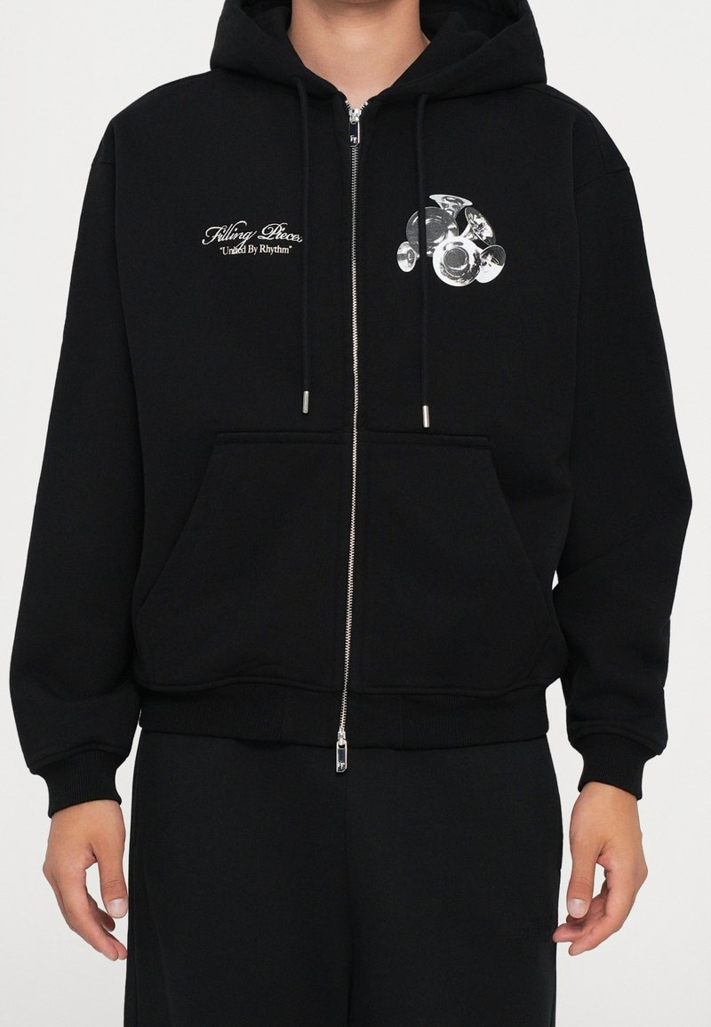 Sweat à capuche noir zippé avec poches frontales, motif graphique blanc d'un groupe de trompettes sur la poitrine gauche et texte « Filling Pieces United By Rhythm » sur la poitrine droite.