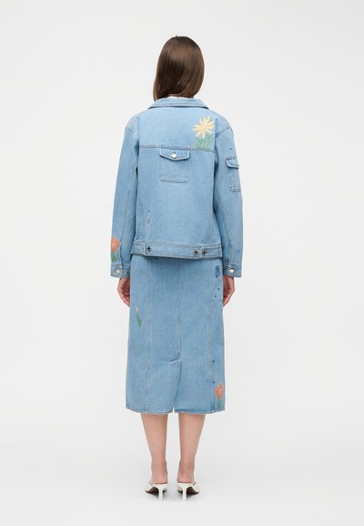 Kvinde iført lyseblå denimjakke og midinederdel med farverig blomsterbroderi, stående med ryggen mod, iført hvide mulehøjhælede sko.