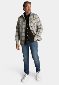 Geruite flanellen shirt in beige en blauw, met twee borstzakken; gecombineerd met donkere spijkerbroeken en witte sneakers, tegen een effen achtergrond.