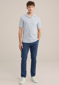 Camisa polo de manga corta azul claro con un patrón geométrico, combinada con chinos azul marino y zapatillas blancas, de pie frente a un fondo neutro.