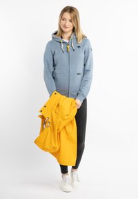 Blaue Zip-Up-Hoodie mit Kordeln und Logo, kombiniert mit einer leuchtend gelben Regenjacke, die in der Hand gehalten wird, schwarzen Hosen und weißen Sneakers.