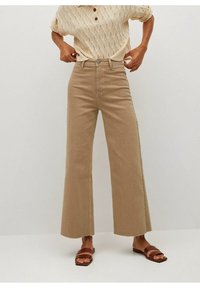 Personne portant un pantalon large taille haute beige, un haut en tricot crème à manches courtes et des sandales plates brunes, ajustant la ceinture contre un fond uni.