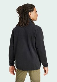Black fleece jacket with a high collar, textured surface, and relaxed fit, shown from the back, paired with light green pants.