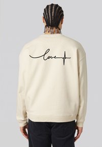Felpa color crema con una vestibilità rilassata, caratterizzata da testo nero "love" con una linea del battito cardiaco sul retro. Tessuto texturizzato, polsini a coste.