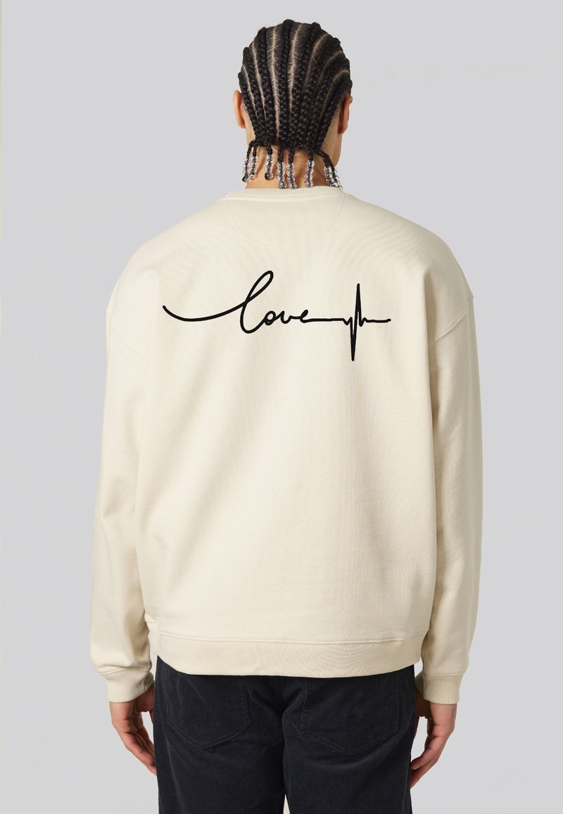 Felpa color crema con una vestibilità rilassata, caratterizzata da testo nero "love" con una linea del battito cardiaco sul retro. Tessuto texturizzato, polsini a coste.