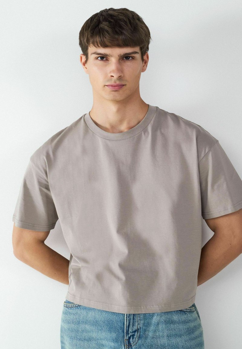 Jasnoszara koszulka z krótkim rękawem wykonana z miękkiego materiału, z okrągłym dekoltem i skróconym fasonem. Noszona z niebieskimi dżinsami.