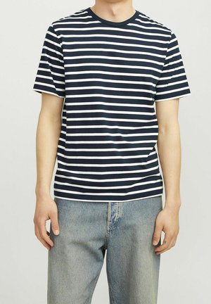 Personne portant un t-shirt à manches courtes à rayures horizontales bleu marine et blanc et un jean en denim bleu clair, debout devant un fond uni.