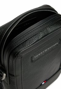 Bolso cruzado de cuero negro con cierre de cremallera, superficie texturizada, parche con logo en el frente y una franja de acento en rojo, blanco y azul.