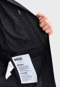 Interno di una giacca nera caratterizzato da una texture di tessuto liscia, una tasca laterale con zip e un'etichetta bianca con testo nero.