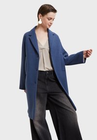 Modrý kabát z vlnenej zmesi s jednoradovým zapínaním, klopovými goliermi, prednými vreckami a voľným strihom, skombinovaný s svetlým topom a širokými džínami.