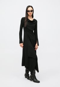 Vestido preto de manga comprida com decote drapeado, bainha assimétrica e fendas laterais. Usado com óculos de sol pretos e botas de couro até ao joelho.