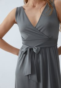 Harmaa kietaisumekko, jossa on syvä V-aukko, istuva yläosa ja vyötäröllä solmittava yksityiskohta. Valmistettu pehmeästä, sileästä kankaasta, jossa laskeutuva hame.