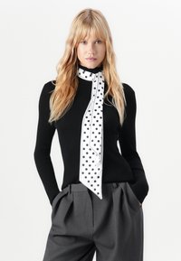 Pull noir à côtes avec des manches longues associé à un pantalon gris à plis. Écharpe blanche à pois noirs nouée autour du cou.