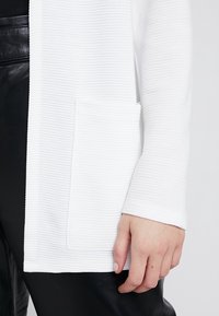 Cardigan bianco con texture e sottile motivo a coste, dotato di una tasca frontale e design aperto. Abbinato a pantaloni in ecopelle neri.