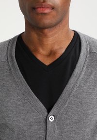 Grå cardigan i mjukt material, med V-ringning, knäppning och bärs över en svart T-shirt.