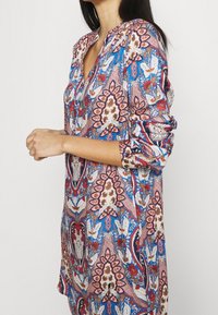 Nainen pitkähihaisessa tunikassa, jossa on sinisiä, vaaleanpunaisia ja valkoisia kukka- ja paisley-kuvioita, seisomassa yksiväristä taustaa vasten.