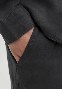 Détail d'une chemise en lin noir montrant un revers de manche roulé, une main atteignant une poche latérale avec une texture et des coutures visibles sur le tissu.