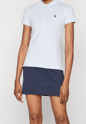 Kvinde iført en lyseblå poloshirt og en mørkeblå nederdel, stående foran en ensfarvet lys baggrund.