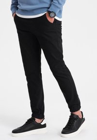 Svarta joggingsbyxor med en smal passform, elastiska muddar och sidofickor, kombinerade med svartvita sneakers, mot en neutral bakgrund.