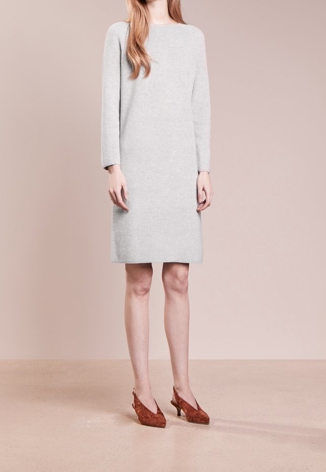 Femme portant une robe gris clair à manches longues et longueur genoux ainsi que des chaussures marron à bout pointu et talon bas sur un fond neutre.