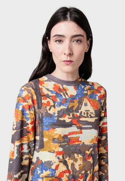 Woman with long dark hair wearing a colorful long-sleeve shirt featuring an abstract landscape with trees and a house.