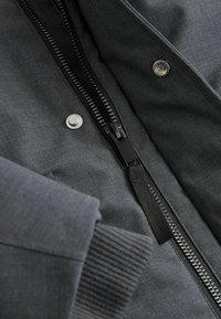 Nahaufnahme eines dunkelgrauen Jackenärmel mit geripptem Bündchen, schwarzem Reißverschluss und zwei silbernen Druckknöpfen auf dem Stoff.