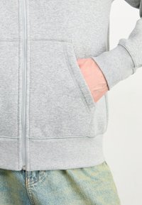 Felpa grigia con zip realizzata in un tessuto morbido, dotata di una tasca frontale, polsini a costine e un design semplice. Indossata con jeans azzurri.