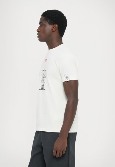 Camiseta blanca de algodón con gráficos impresos en negro y rojo. Mangas cortas con una etiqueta de parche en el brazo izquierdo. Corte clásico.