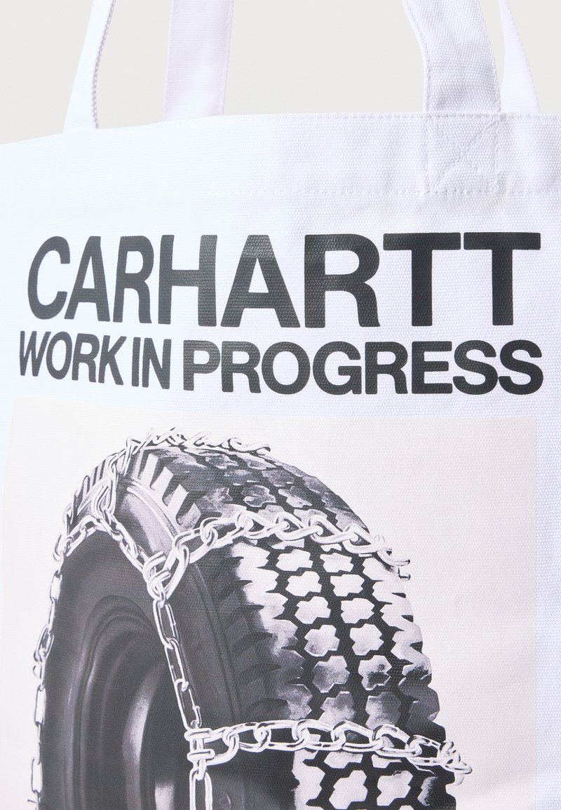 Sac fourre-tout blanc Carhartt avec texte noir "CARHARTT WORK IN PROGRESS" et illustration d'un pneu entouré de chaînes.
