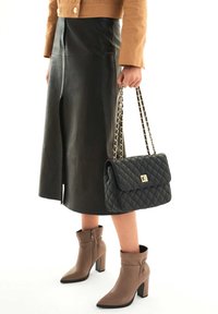 Borsa a tracolla nera trapuntata con una chiusura dorata, indossata con una giacca corta marrone, una gonna in pelle nera e stivaletti taupe con tacco spesso.