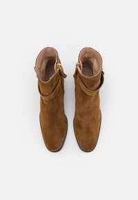 Hnědé semišové ankle boots s hladkým vnitřním podšívkou, zlatými kovovými akcenty a designem s ohnutým límcem pro přidaný detail.