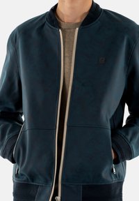 Chaqueta bomber de color navy, hecha de material sintético liso, con cremallera metálica, puños acanalados y bolsillos laterales. Pequeño parche con logo en el pecho.