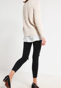 Beige neulepusero, jossa on teksturoitu kuvio, kerroksittain valkoisen paidan päällä, yhdistetty mustiin skinny-farkkuihin, joissa on vetoketjut nilkoissa.