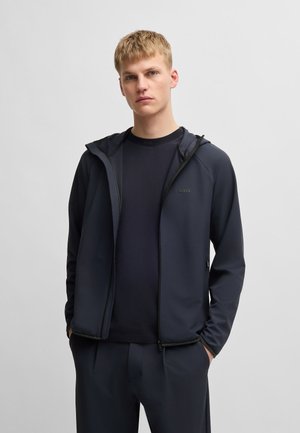 Junger Mann trägt eine dunkelblaue Kapuzenjacke mit Reißverschluss über einem marineblauen Rundhalsausschnitt, steht mit den Händen in den Taschen vor einem schlichten grauen Hintergrund.