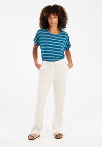 Mėlynos ir žalios juostelės trumpas rankovės marškiniai su laisvu siluetu, derinami su grietinėlės spalvos siaurėjančiais šortais ir smėlio spalvos šlepetėmis. Lygus paviršius, kasdieniškas dizainas.