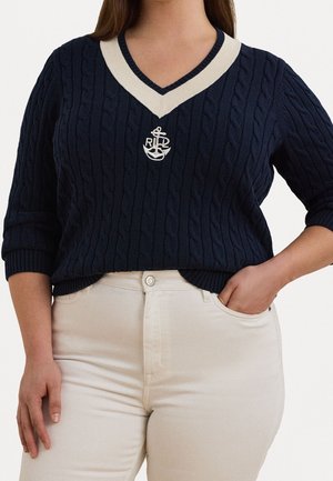 Kvinde iført navyblå kabelstrikket V-hals sweater med hvidt ankerlogo og cremefarvede bukser med højtaljet pasform, den ene hånd i lommen.
