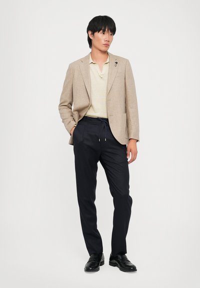 Beige strukturierter Blazer über einem hellgelben Poloshirt, kombiniert mit navyblauen tapered Hosen und schwarzen Slippern. Stehende Pose vor einem weißen Hintergrund.