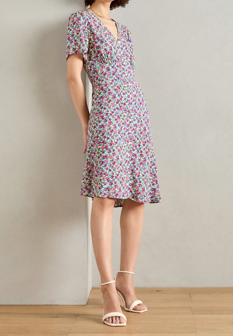 Femme portant une robe fleurie mi-longue à manches courtes et col en V, associée à des sandales à talons hauts beige, debout à l'intérieur.