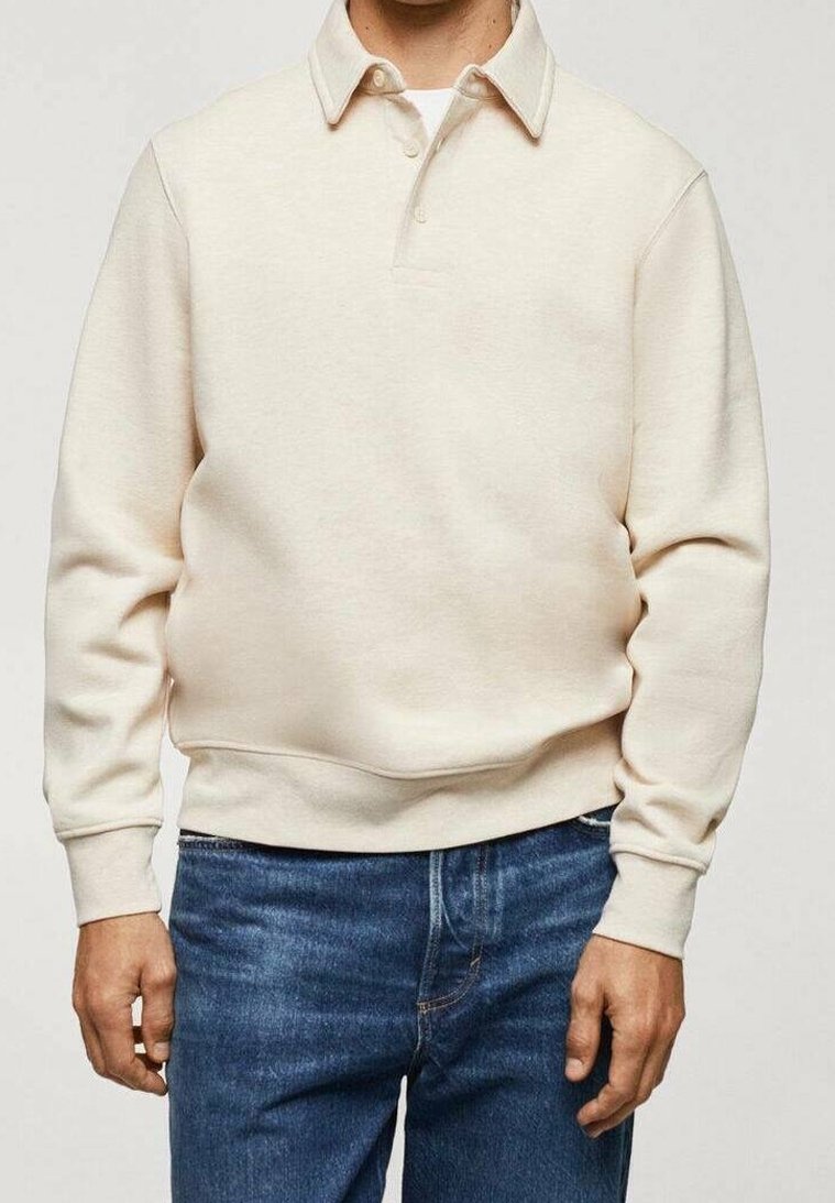 Suéter beige de punto con cuello tipo polo, botones frontales y mangas largas. Confeccionado en una suave tela texturizada, combinado con pantalones vaqueros azules.