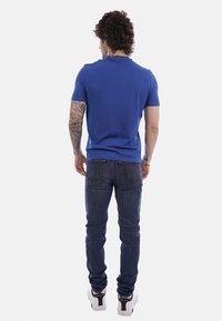 Homme aux cheveux bouclés et tatouages sur le bras gauche, portant un t-shirt bleu, un jean foncé et des baskets blanches, debout de dos sur un fond blanc.