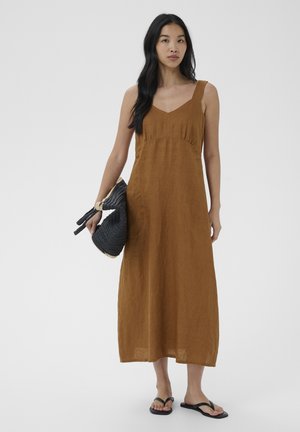 Femme aux longs cheveux noirs portant une robe midi marron sans manches et des tongs noires, tenant un sac tressé foncé dans sa main gauche.