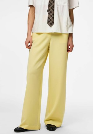 Une personne porte un pantalon ample jaune pâle, une chemise blanche à manches courtes, une cravate à carreaux marron et des chaussures noires sur un fond blanc.