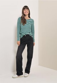 Groen en wit gestreepte langwerpige blouse met een relaxte pasvorm, gecombineerd met zwarte wijde broekspijpen en witte sneakers op een neutrale achtergrond.