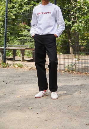 Person trägt weißen Pullover mit rotem "Carhartt"-Logo, schwarze Hose und weiße Turnschuhe, steht auf Kies neben einem Metallzaun im Freien.