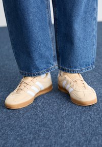 Beige mockasinsneakers med vita ränder, rundad tå och gummisulor, tillsammans med vida blå denimbyxor. Ytan är slät och mjuk.